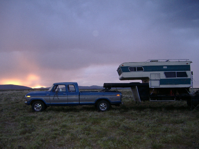 Blue and silver Ford pickup truck towing a camper, side view. Sunset in background