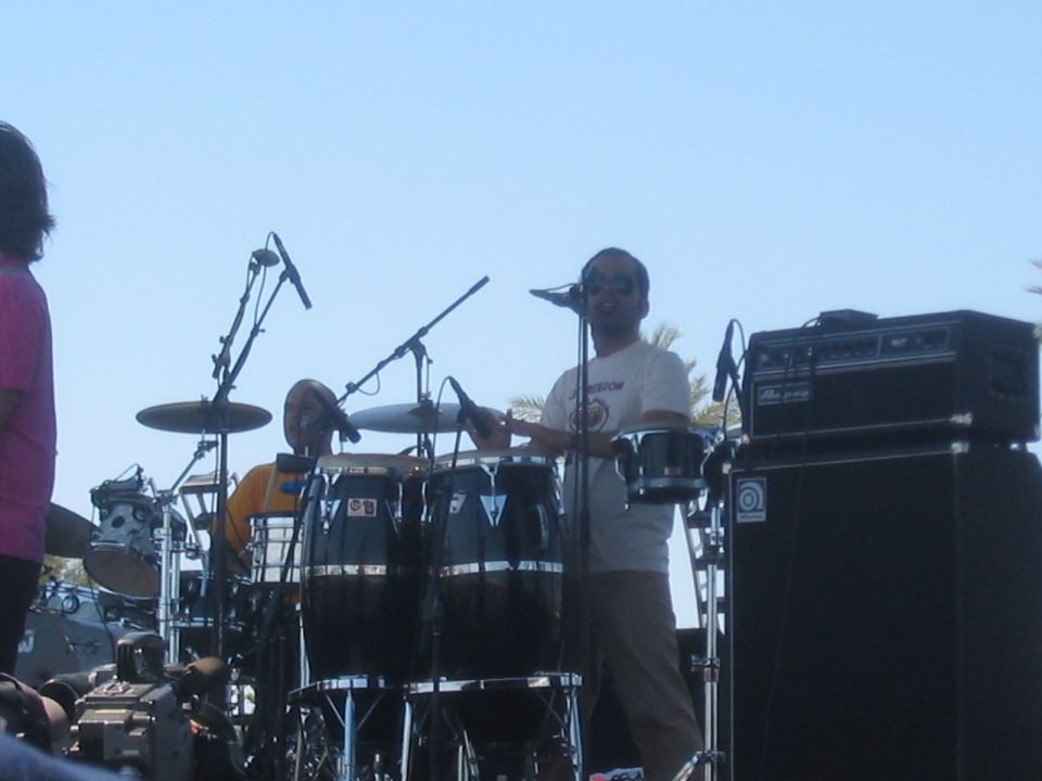 Musicians play drums and congas on an outdoor stage with microphones and amplifiers