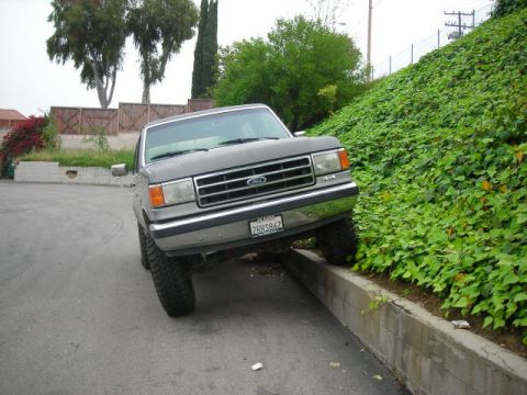 1988 Ford Bronco - This Ford Bronco is parked with its front wheels on a concrete curb next to a grassy hill.