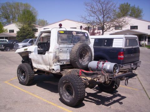 1978 Ford Bronco - Modified pickup truck chassis with spare tire and propane tank, parked behind another truck.