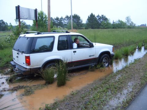 1988 Ford Bronco - A white Ford Explorer is stuck in a muddy ditch off a rural road.