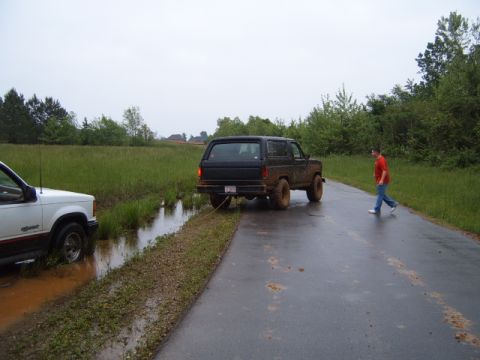 1988 Ford Bronco - A black SUV is stuck in the mud and being towed by a white SUV on a wet road.