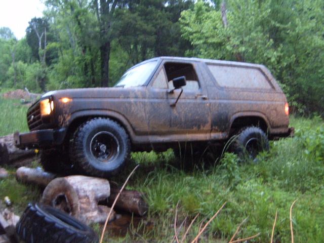 Muddy 1980s Ford Bronco driving over logs and grass