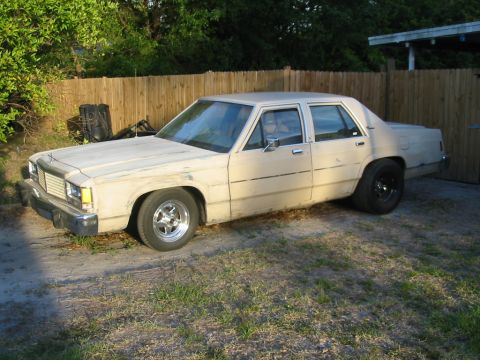 1985 Ford LTD Crown Victoria - A tan sedan with a wooden fence in the background and aftermarket wheels is parked on a gravel surface.