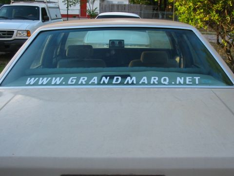 1985 Ford LTD Crown Victoria - The rear of a tan sedan shows a large white sticker on the window, with another vehicle parked nearby.