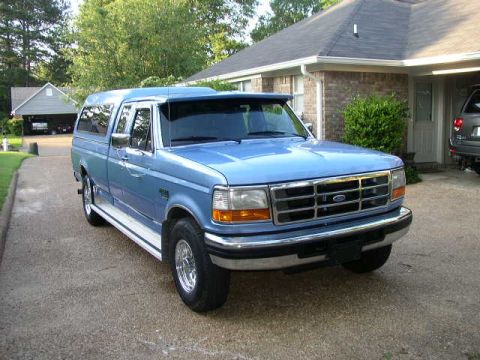 1995 Ford Bronco - A blue pickup truck with a camper shell is parked in front of a brick house.