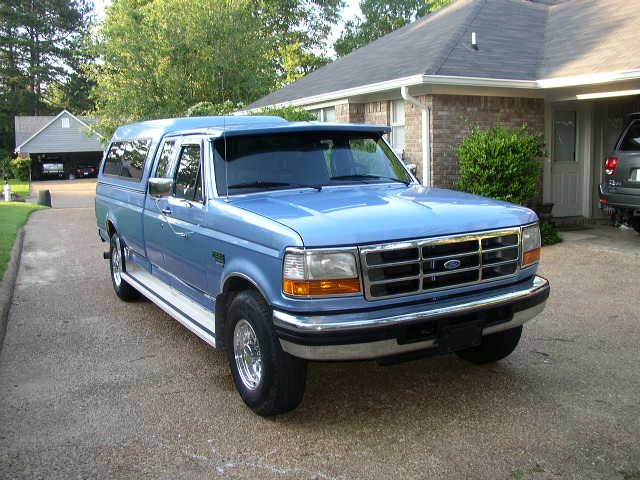 Blue pickup truck with a camper shell parked in a driveway. It has a chrome grille and chrome wheels