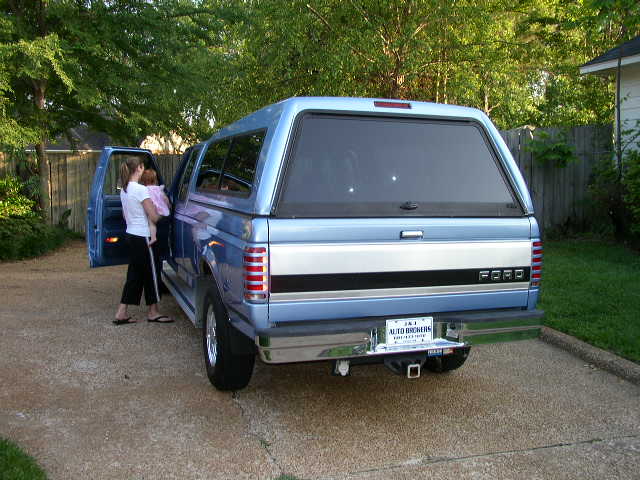 Blue Ford pickup truck with camper shell, woman holding baby near driver's door