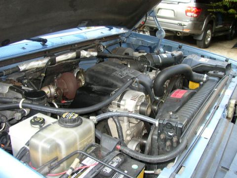 1995 Ford Bronco - The engine bay of a blue vehicle is shown, a turbocharger and other engine components. Another SUV is visible in the background.