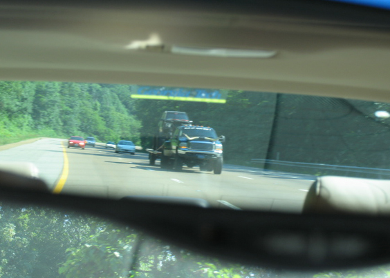 Rear-view mirror shot of a dark pickup towing a loaded flatbed trailer on a highway