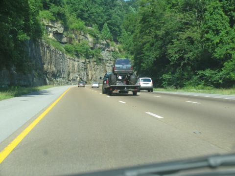 1988 Ford E250 - A dark pickup truck tows a dark SUV on a trailer along a highway with a rocky cliff face and trees.