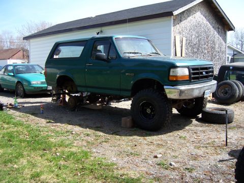 1996 Ford Bronco - A green 1996 Ford Bronco is elevated on jack stands, showing its lifted suspension and off-road tires, with another vehicle parked nearby.