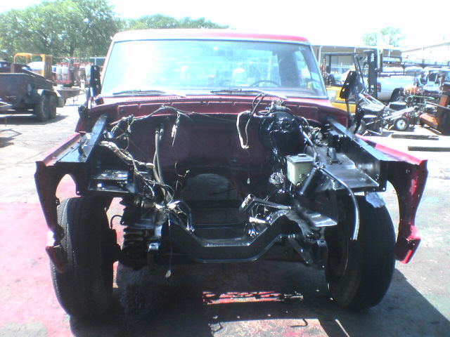 Bare front frame and suspension of a dark red truck in a junkyard, other vehicles in background
