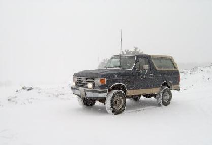 1991 Ford Bronco - A dark blue full-size Ford Bronco with a tan removable rear top sits in deep snow during a whiteout, photographed from the three-quarter fr…