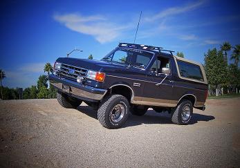 1991 Ford Bronco - Three-quarter front view of a dark blue and tan two-tone full-size Ford Bronco parked on pavement with palm trees and blue sky behind it. I…