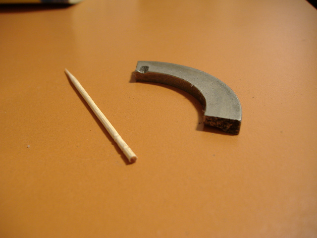Curved metal bearing piece next to a wooden toothpick on an orange surface