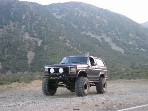 1985 Ford Bronco - Modified 1985 Ford Bronco with large tires and lift kit sits on a dirt road below a mountain landscape.
