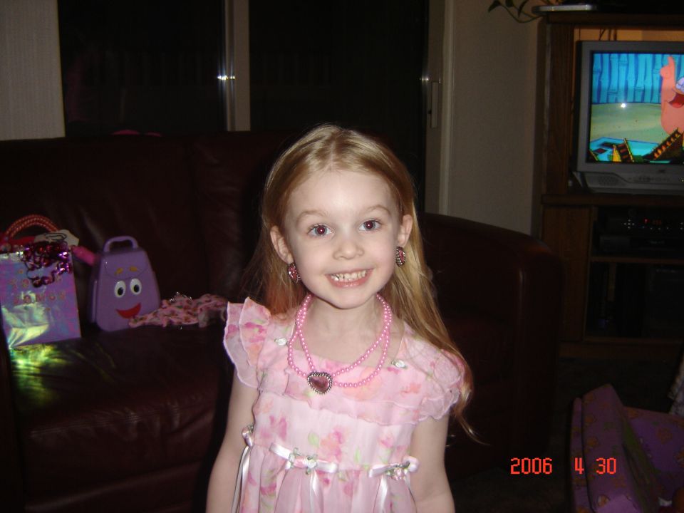 Girl in a pink floral dress and necklace smiles, with a purple toy and gift bags on a leather couch