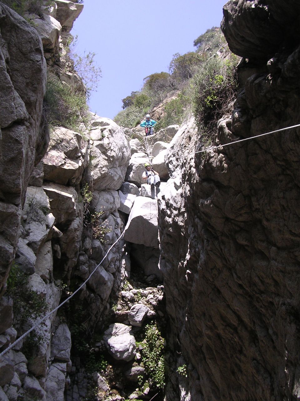 Two people are rappelling down a steep, rocky canyon with ropes and harnesses. Vegetation grows on the canyon walls