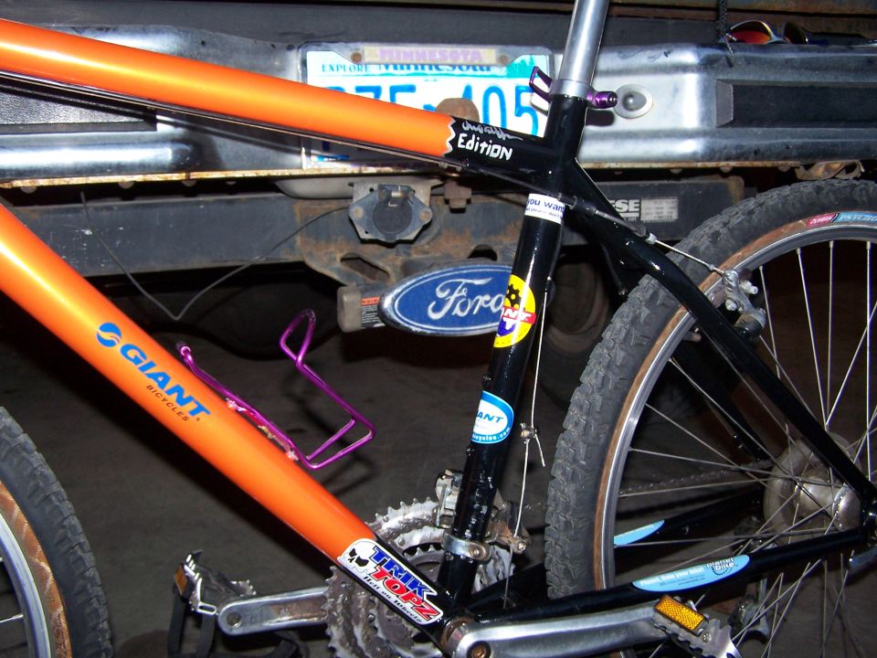 Orange Giant bike leaning against truck bumper with Ford oval and Minnesota plate behind it
