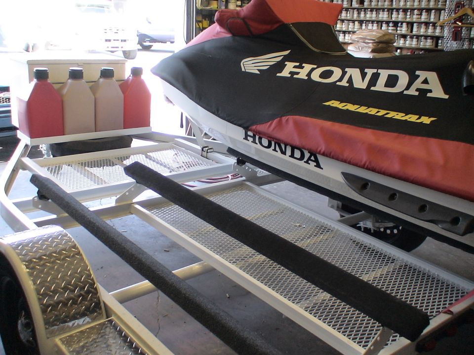 Covered Honda AquaTrax personal watercraft on a white double trailer with fuel jugs in a garage