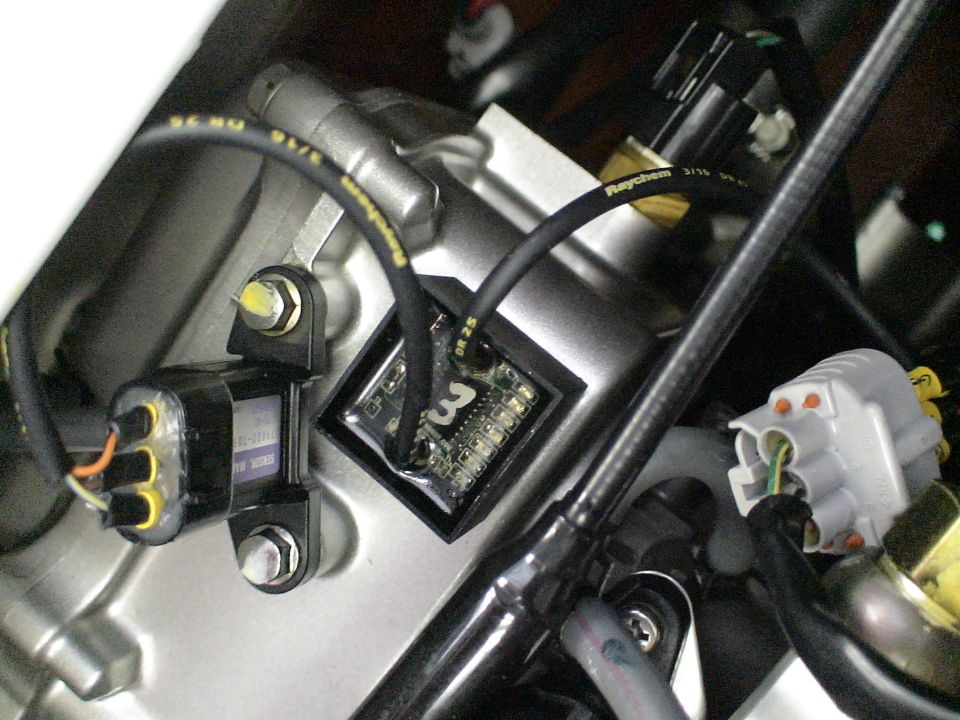 Close-up of engine bay wiring, labeled black cables, a control module and white connectors over silver housing