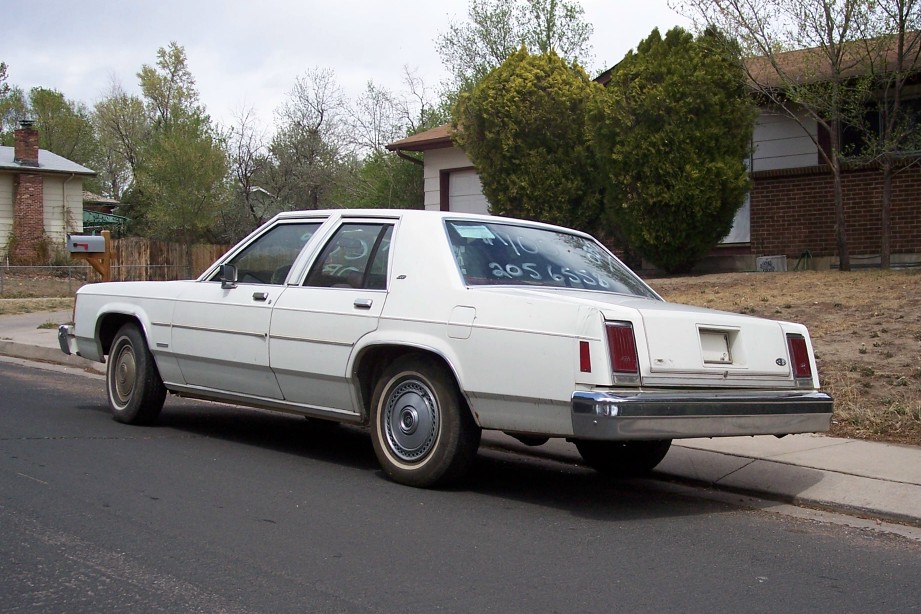 White sedan parked on street, rear view. Numbers written on rear window