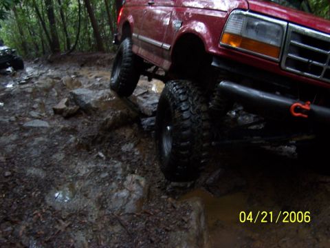 1995 Ford Bronco - Close low-angle shot of a lifted red Ford Bronco flexing on wet rock, with the driver-side front mud-terrain tire stuffed up and the rear w…