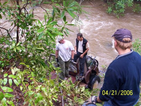 1995 Ford Bronco - A dark red Jeep lies rolled on its side in a swollen muddy river while two men stand on the body and tub working a recovery, with the roll…