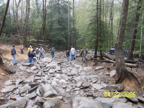 1995 Ford Bronco - A group of wheelers and spectators stands along a wet, rock-strewn creek-bed obstacle in a wooded off-road park, scouting the line up the b…