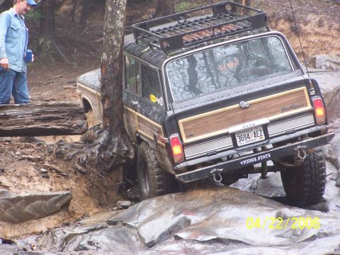 1995 Ford Bronco - Rear three-quarter view of a mud-covered black Jeep Grand Wagoneer with woodgrain side trim, roof rack and steel rear bumper with tow hooks…