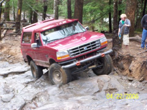 1995 Ford Bronco - A red Ford Bronco with hardtop crawls across a wet rock slab on a wooded trail, leaning hard with its front passenger tire lifted, riding o…