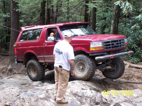 1995 Ford Bronco - A man watches as a red 1995 Ford Bronco tackles a challenging off-road course in a wooded area.