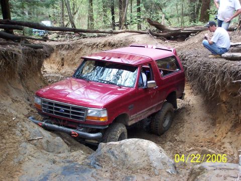 1995 Ford Bronco - Front three-quarter view of a red Ford Bronco with hardtop working through a deeply rutted, eroded dirt trail between clay banks, flexing i…