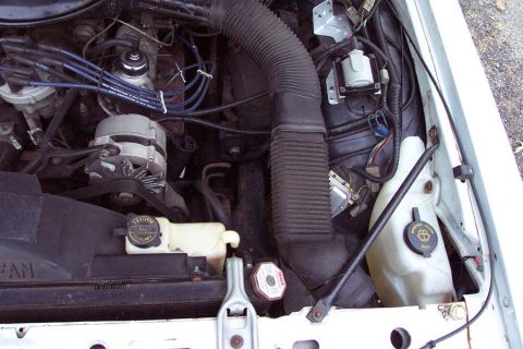 1985 Ford LTD Crown Victoria - Engine bay of a white car looking down the passenger side, with a large black corrugated air intake duct running from the fender to the thr…
