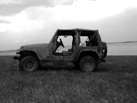 1998 Jeep TJ - A muddy 1998 Jeep TJ is seen from the side, parked in a field with a body of water in the background.