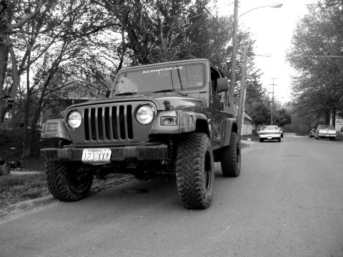 1998 Jeep TJ - A dark 1998 Jeep TJ is parked on a residential street, showing off its large tires and a club sticker on the windshield.