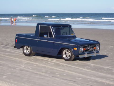 1987 Suzuki Samurai - A blue pickup truck with custom wheels sits on the sand near the ocean waves.