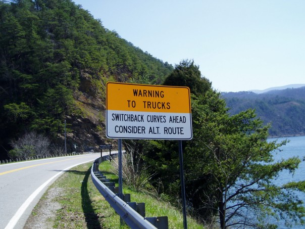 Roadside sign warns trucks of switchback curves ahead and suggests an alternate route. Mountainous terrain and water are visible