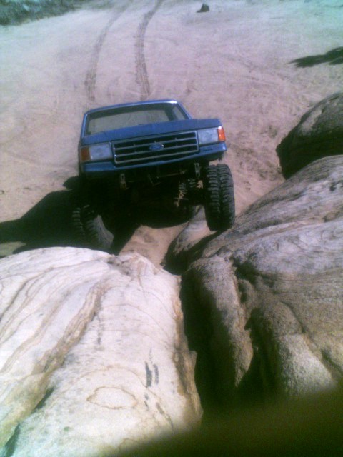 1989 Ford Bronco - A blue Ford truck is shown off-roading through a rocky landscape, with tire tracks visible in the dirt.