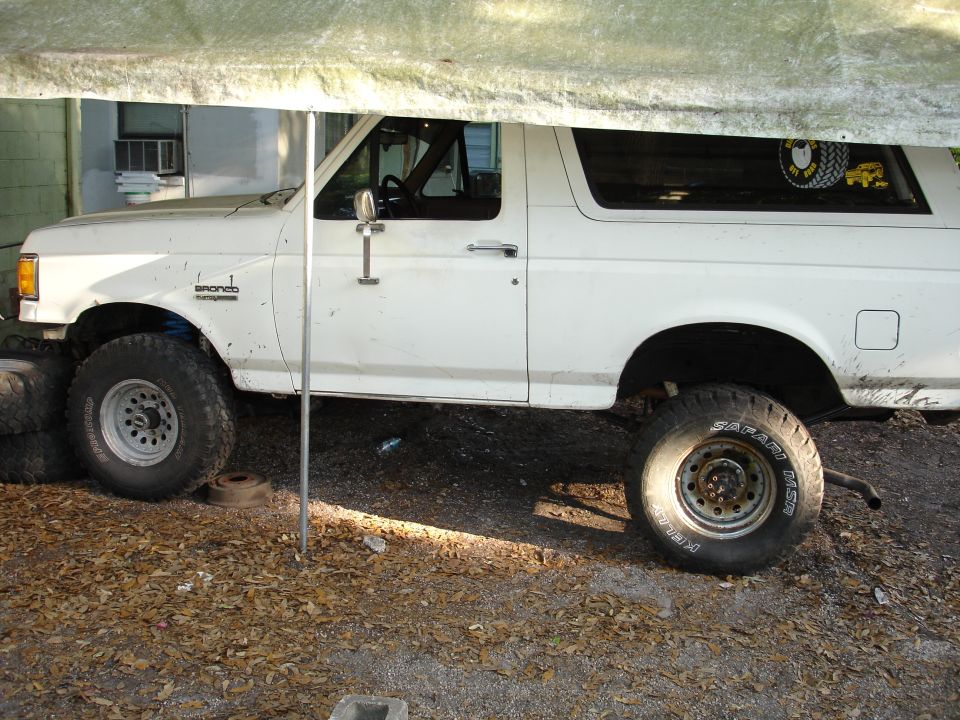 White Ford Bronco with aftermarket wheels and tires, parked under a tarp