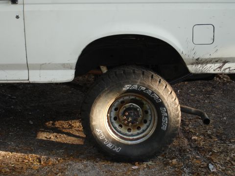 1989 Ford Bronco - Close-up of the rear wheel and fender of a white pickup truck, showing off-road tires and mud.