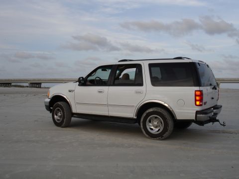 1989 Ford Bronco - This white SUV, likely a 1997-2002 Ford Expedition, is parked on a sandy beach with a bridge visible in the distance.