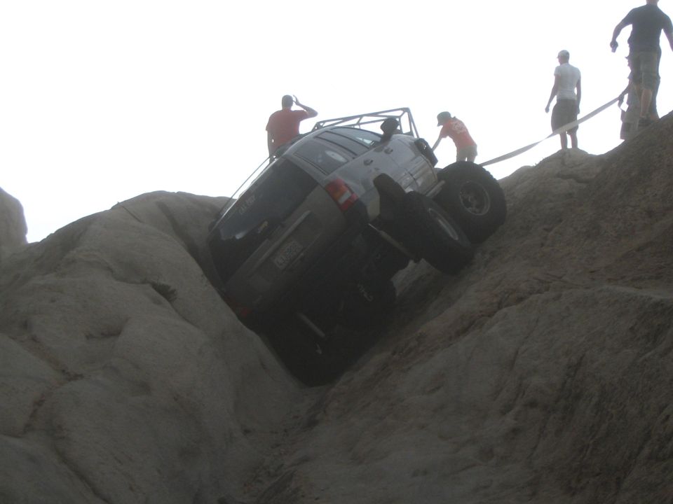 Silver Jeep Grand Cherokee tilted on its side on a rocky incline, with people observing