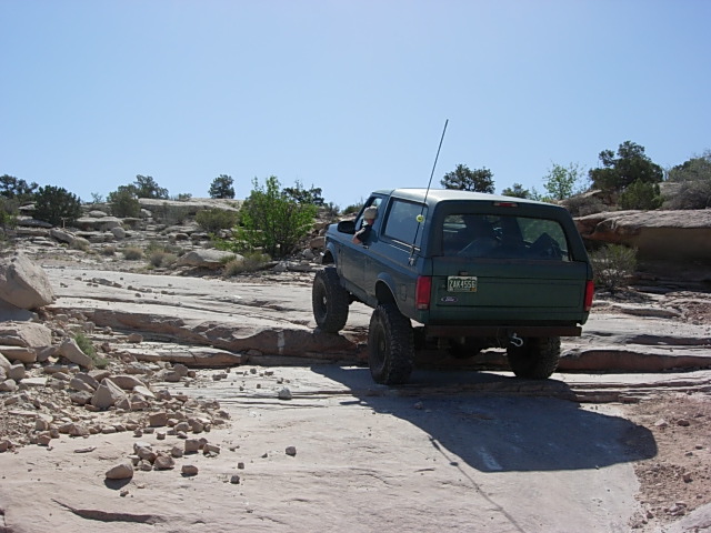 Green 1987 Ford Bronco off-roading on rocky terrain, rear view