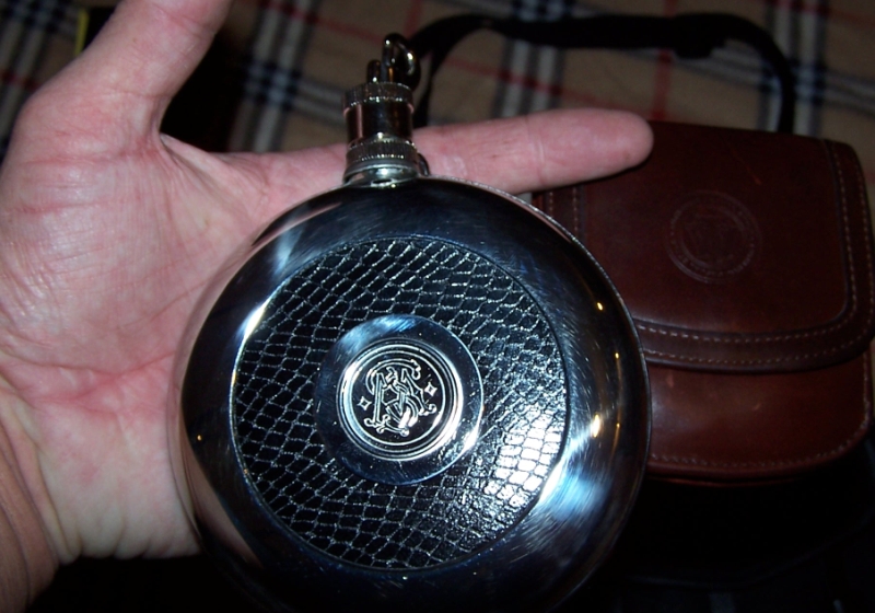 Hand holding round metal flask with textured pattern and emblem, next to brown leather pouch