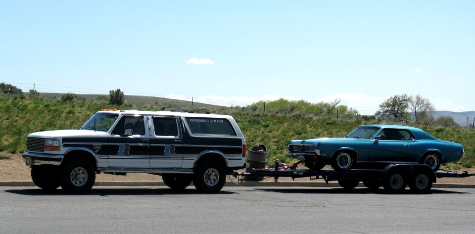 White four-door Ford Bronco towing teal Mercury Cougar on open trailer, side view