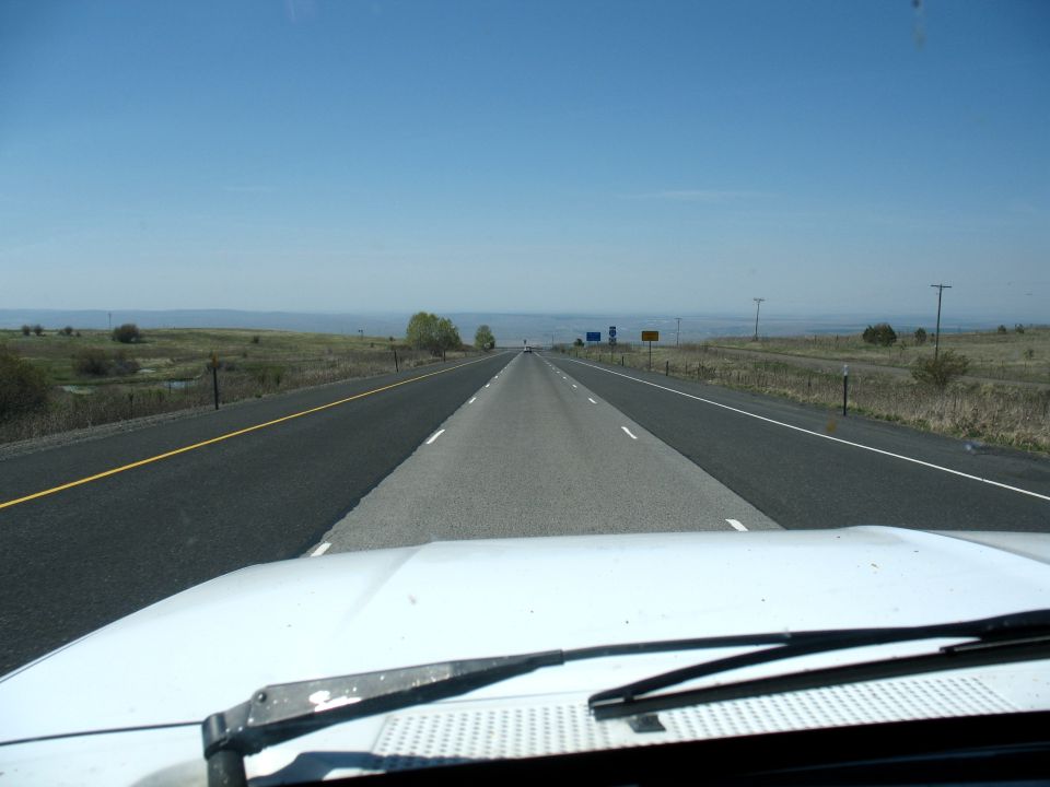 POV over a white vehicle hood down an empty two-lane rural highway under blue sky