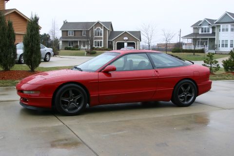 1993 Ford Probe - Side profile of a red Ford Probe on a wet suburban driveway, wearing dark gunmetal five-spoke Cobra R-style wheels and a small 'GT' graphic…