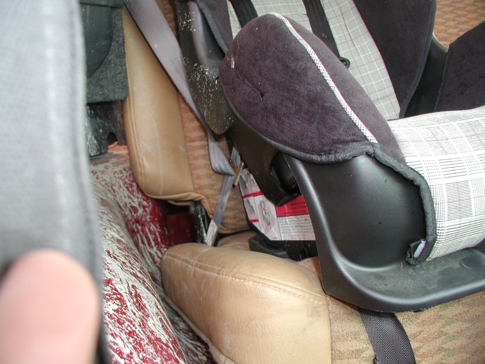 Child car seat installed on tan leather seats, with red and white splatters on the floor